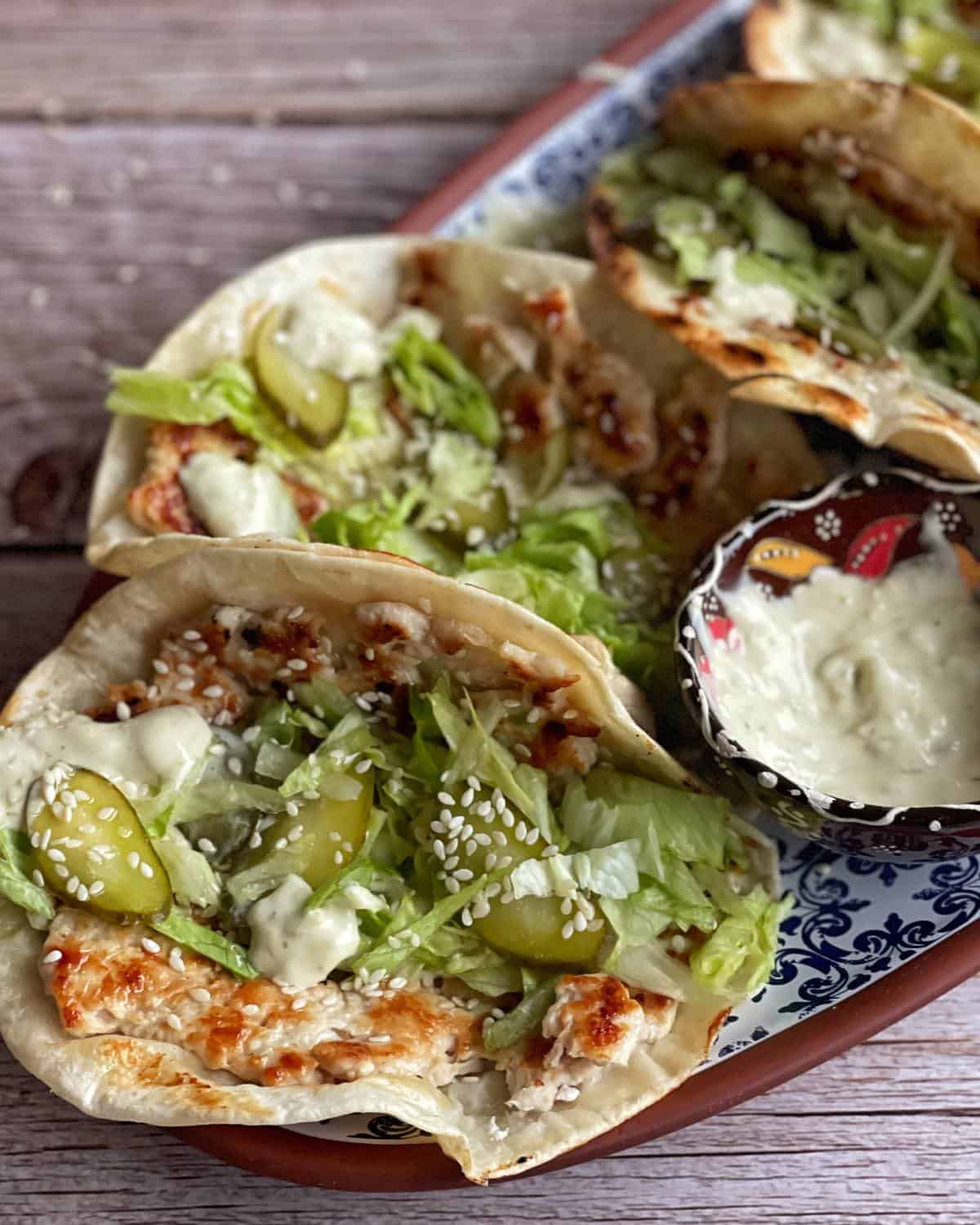 Three soft tacos filled with grilled chicken, lettuce, pickles, sesame seeds, and a creamy white sauce are served on a patterned tray with a small bowl of extra sauce.