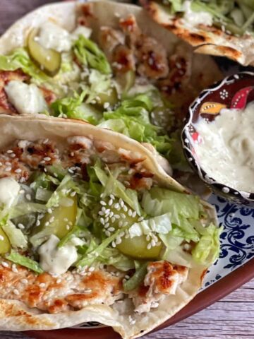 Soft flatbreads topped with grilled chicken, lettuce, pickles, sesame seeds, and white sauce, served on a plate with an extra bowl of sauce on the side.