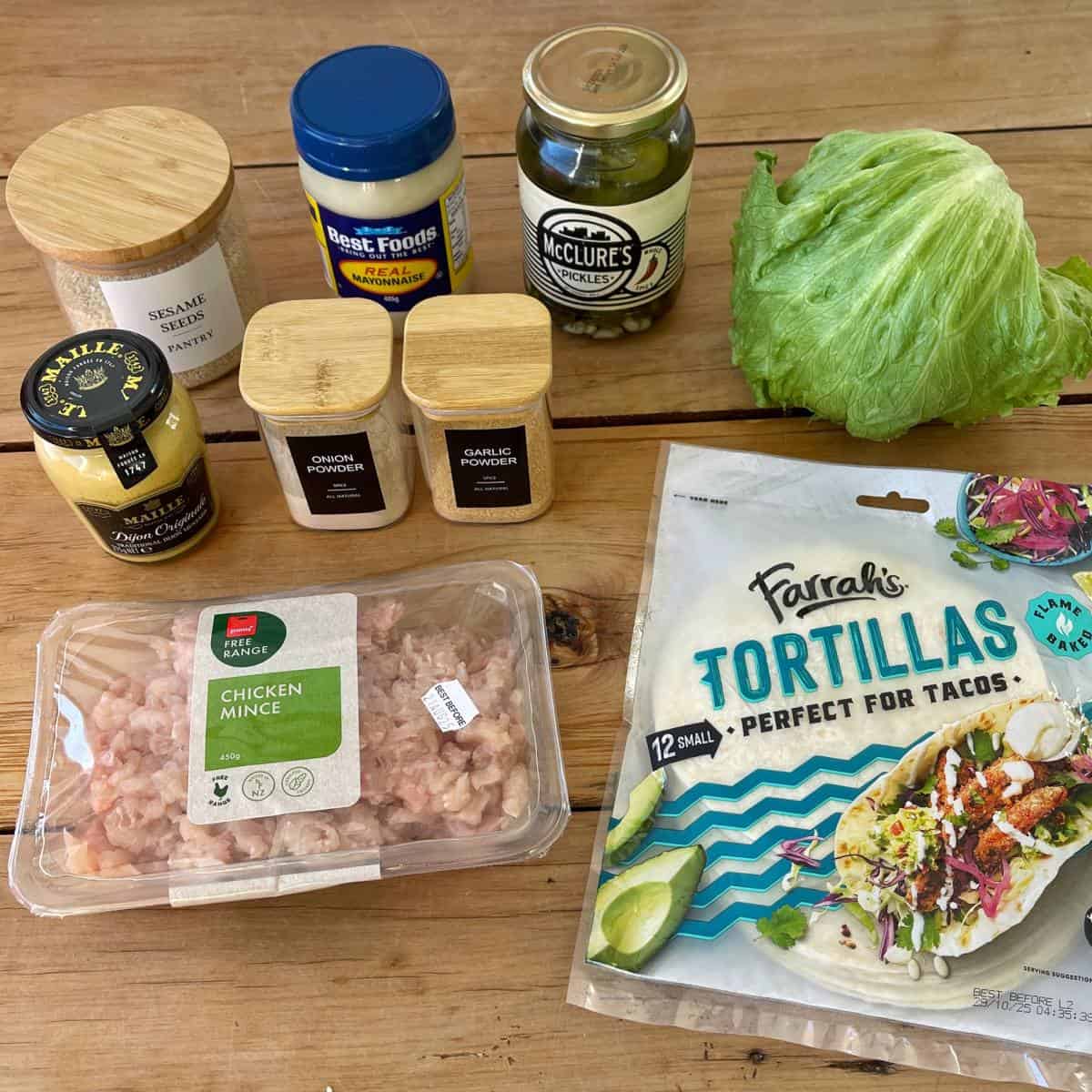 Homemade McChicken Tacos A wooden table displays chicken mince, tortillas, lettuce, pickles, mayonnaise, mustard, sesame seeds, onion powder, and garlic powder.