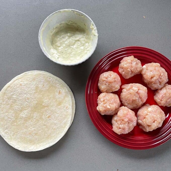 Homemade McChicken Tacos A red plate with raw chicken meatballs, a stack of tortillas, and a bowl of creamy sauce on a gray countertop.