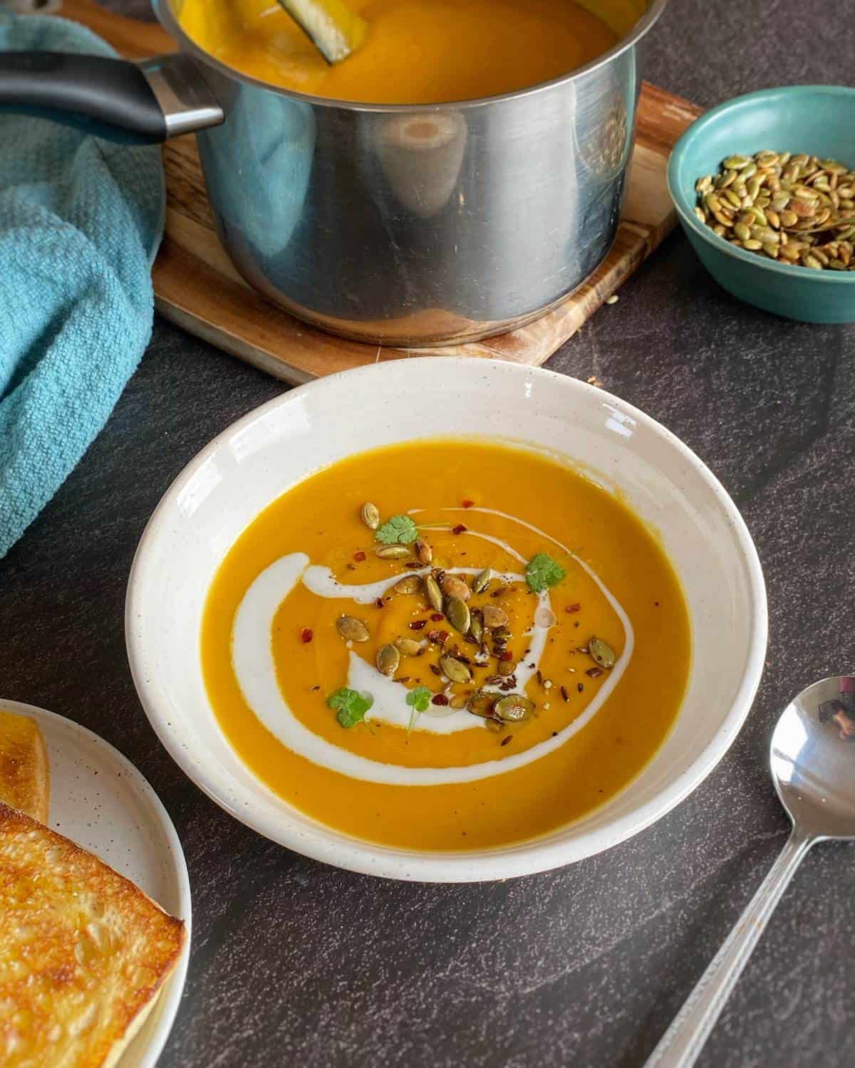 A bowl of orange soup garnished with cream, herbs, and seeds sits on a dark table next to a spoon, toasted bread, and a pot of soup.