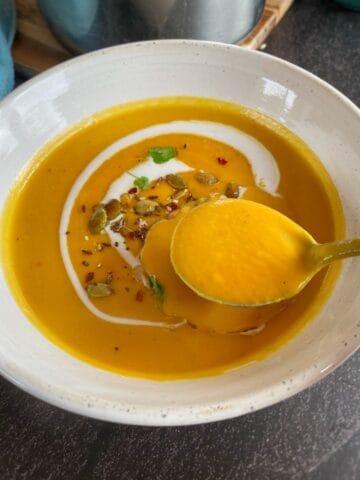 Bowl of orange soup garnished with seeds, herbs, and a swirl of cream, with a spoon lifting some soup.