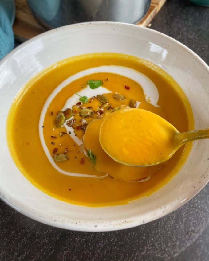 Creamy Coconut and Kumara Soup Bowl of orange soup garnished with seeds, herbs, and a swirl of cream, with a spoon lifting some soup.