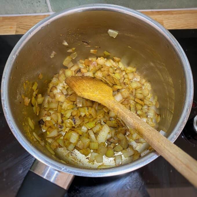 Creamy Coconut and Kumara Soup Chopped onions are being sautéed in a stainless steel pot with a wooden spoon resting inside.