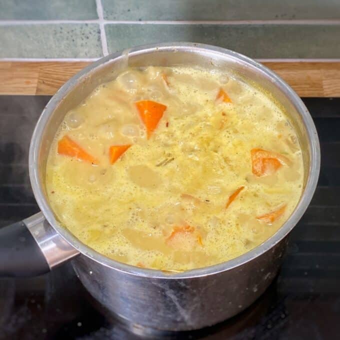 Creamy Coconut and Kumara Soup A metal pot with yellow soup containing orange carrot chunks is boiling on a black stovetop, with green tiles and a wooden countertop in the background.