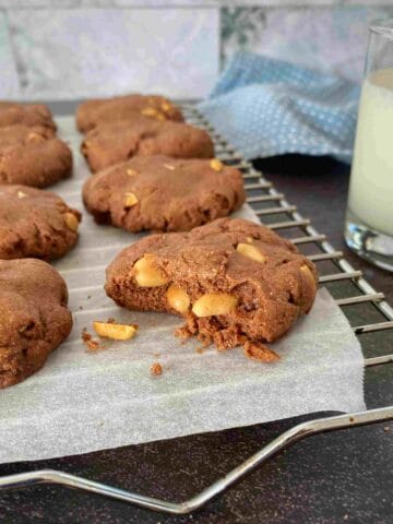 Chocolate cookies with peanuts are placed on a cooling rack lined with parchment paper, next to a glass of milk and a blue cloth in the background.