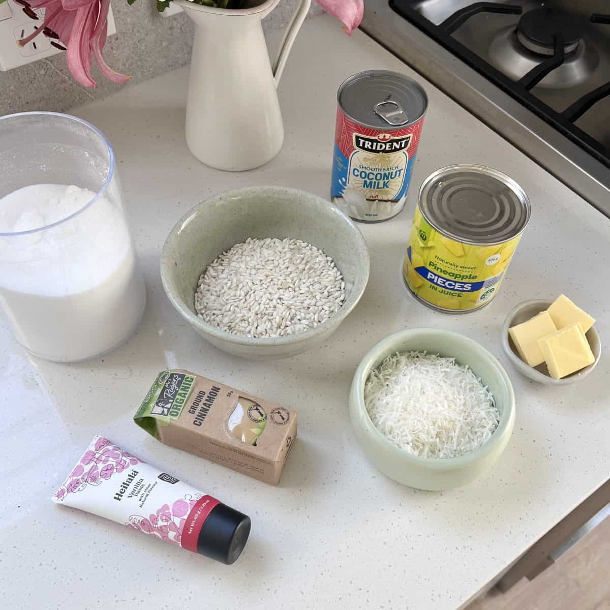 Piña Colada Rice Pudding Ingredients for a recipe on a kitchen counter: sugar, coconut milk, canned pineapple, rice, desiccated coconut, butter, cinnamon, and vanilla extract.