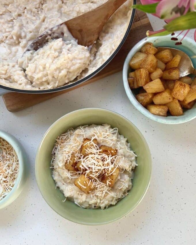 Piña Colada Rice Pudding A bowl of rice pudding topped with caramelized pineapple and shredded coconut sits on a counter next to a pan of rice pudding and a bowl of caramelized pineapple.
