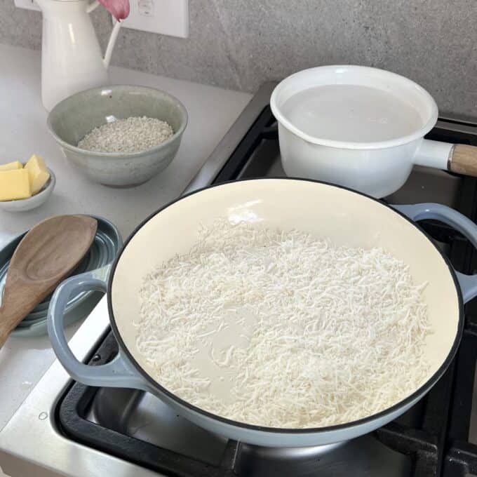 Piña Colada Rice Pudding Dry shredded coconut toasting in a white pan on a stove, with a bowl of rice, a pot of water, butter, and a wooden spoon nearby on a kitchen counter.