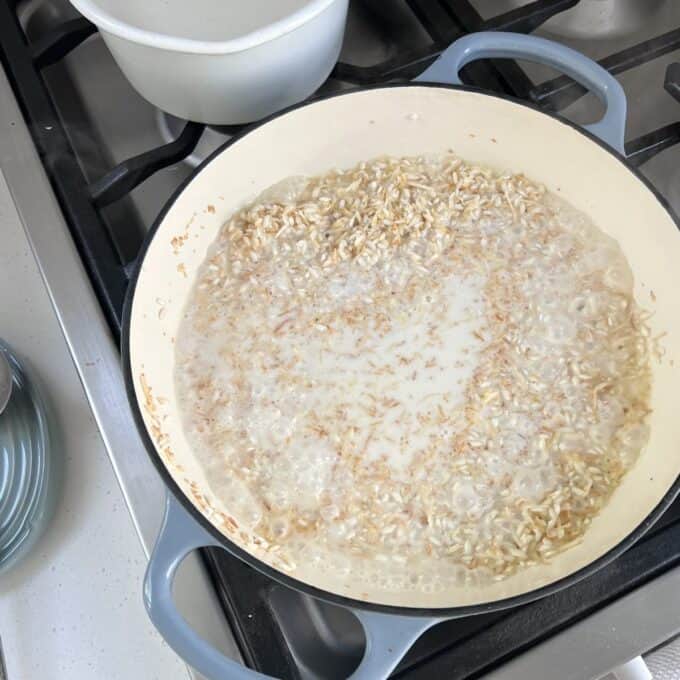 Piña Colada Rice Pudding A pot of oatmeal is simmering on a stove, with bubbles forming on the surface and a white bowl in the background.
