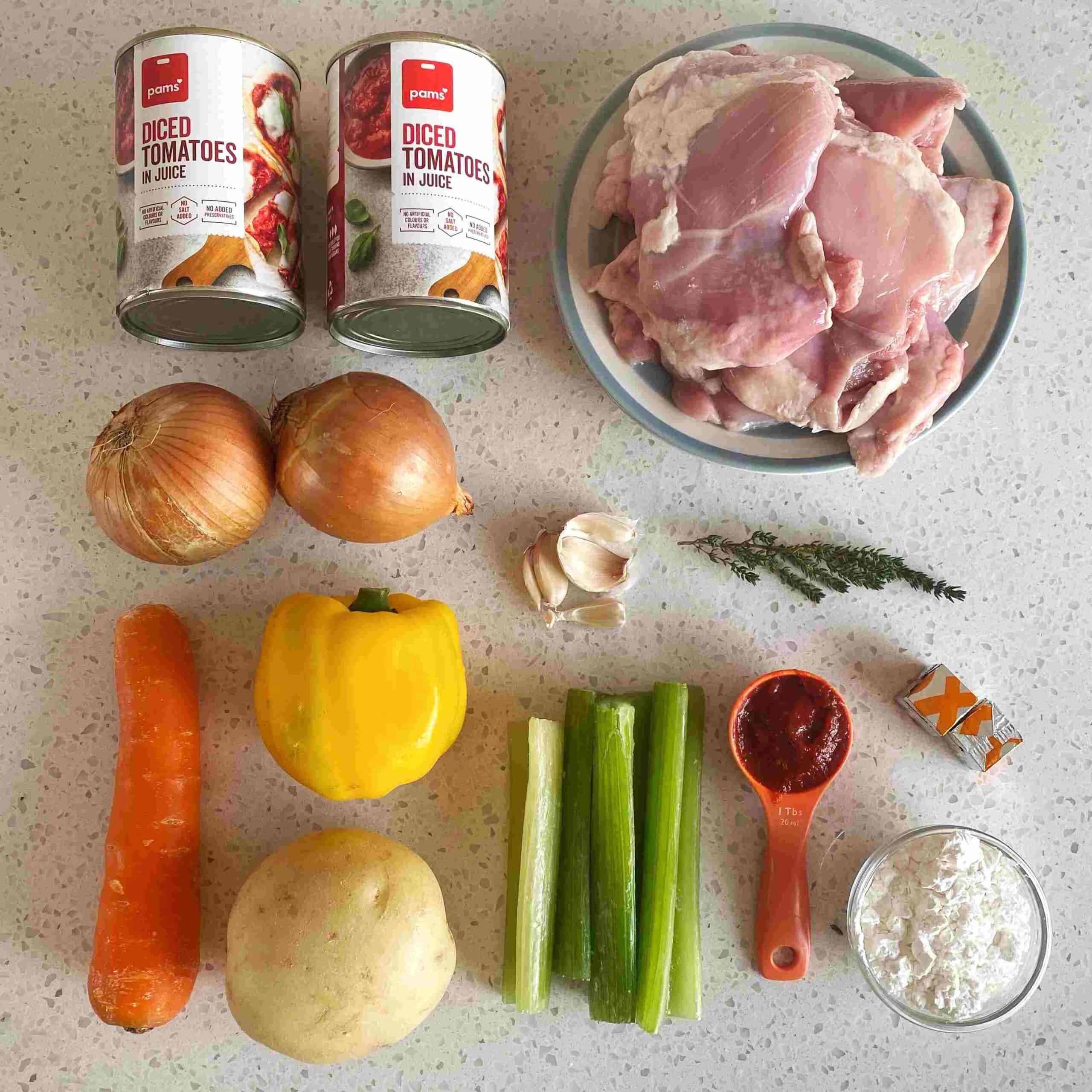 Slow Cooker Tomato Chicken Thigh Stew Ingredients for a recipe arranged on a countertop, including chicken thighs, canned diced tomatoes, onions, carrot, potato, celery, yellow bell pepper, garlic, herbs, stock cube, tomato paste, and flour.