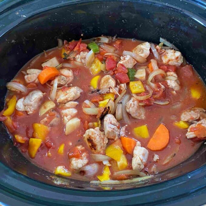 Slow Cooker Tomato Chicken Thigh Stew A slow cooker filled with a stew containing chicken pieces, diced tomatoes, carrots, onions, bell peppers, and celery in a red broth.