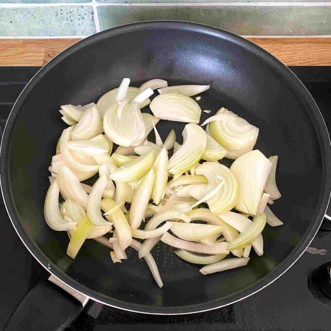 Slow Cooker Tomato Chicken Thigh Stew Sliced onions cooking in a black frying pan on a stovetop.