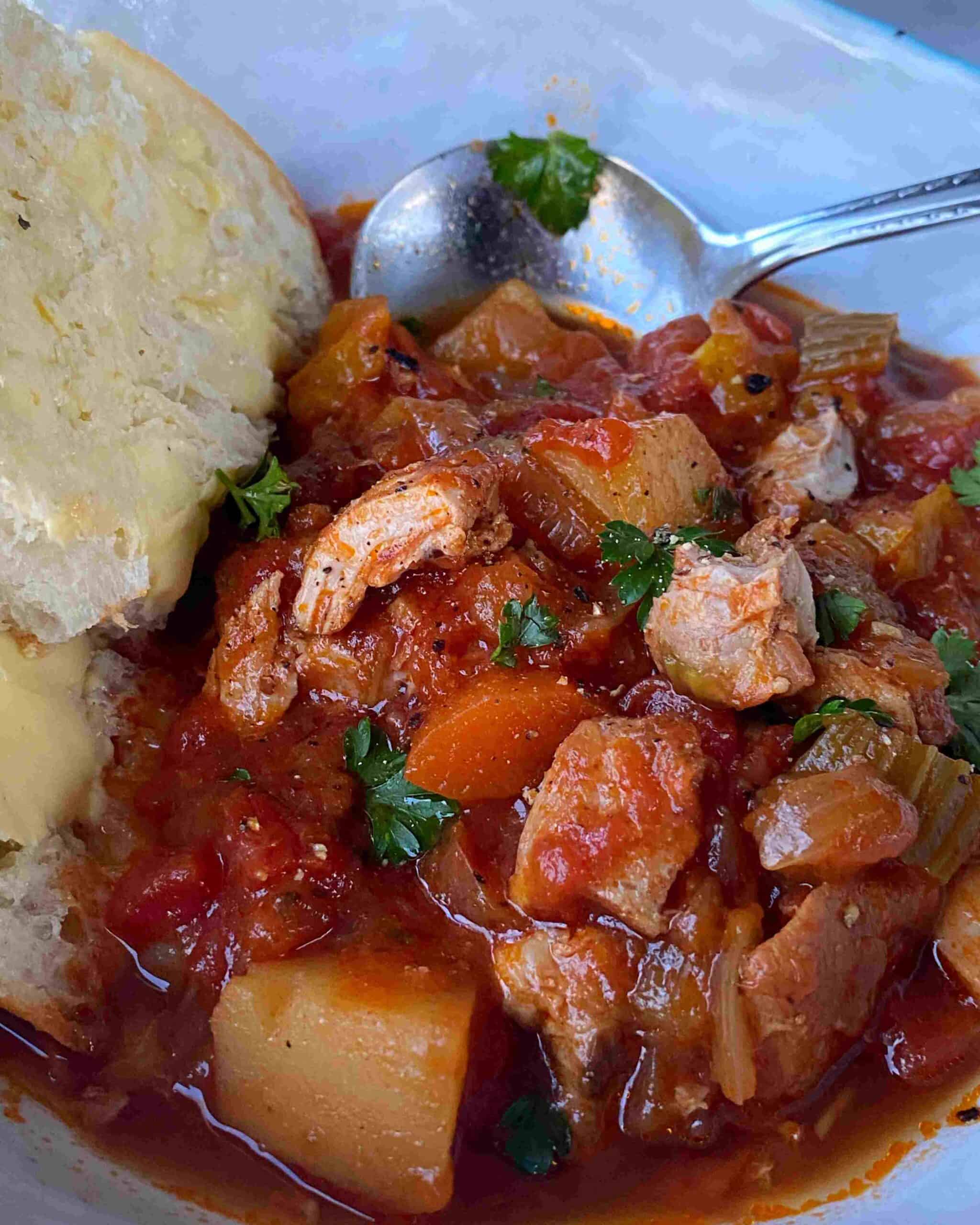 Slow Cooker Tomato Chicken Thigh Stew A close-up of a bowl of chunky chicken and vegetable stew in tomato sauce, garnished with parsley, served with a slice of buttered bread and a spoon.