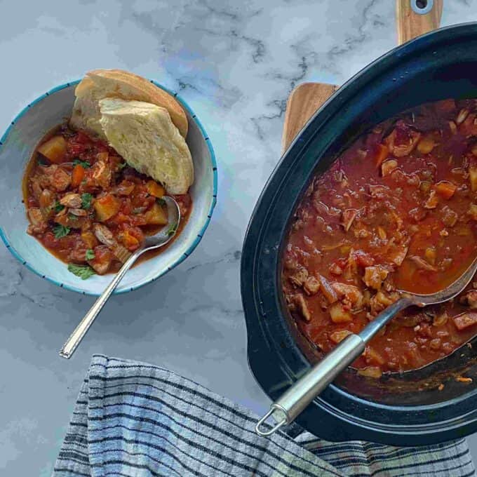 Slow Cooker Tomato Chicken Thigh Stew A bowl of chunky stew with vegetables and bread beside a slow cooker filled with the same stew, set on a marble surface with a striped cloth.