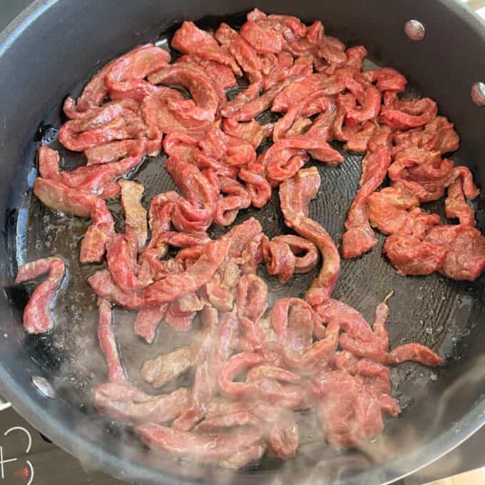 Beef and Broccoli Fried Rice Sliced raw beef strips cooking in a black skillet on a stovetop, with some steam rising from the pan.