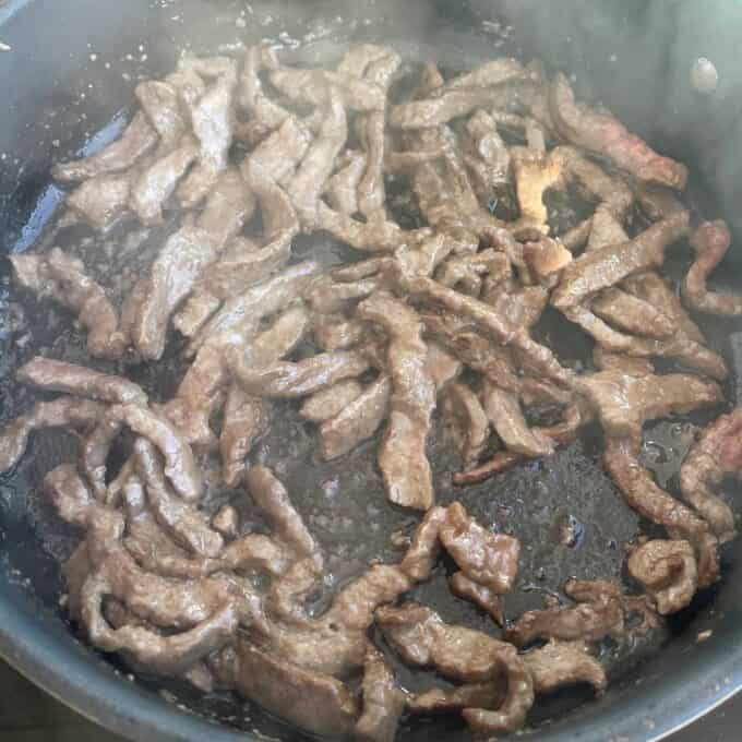 Beef and Broccoli Fried Rice Strips of beef cooking in a frying pan, with visible browning and some oil or liquid in the pan.