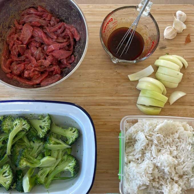 Beef and Broccoli Fried Rice Ingredients for a meal on a wooden surface: sliced raw beef, marinade in a measuring cup, chopped onions, broccoli florets, garlic cloves, and cooked white rice.