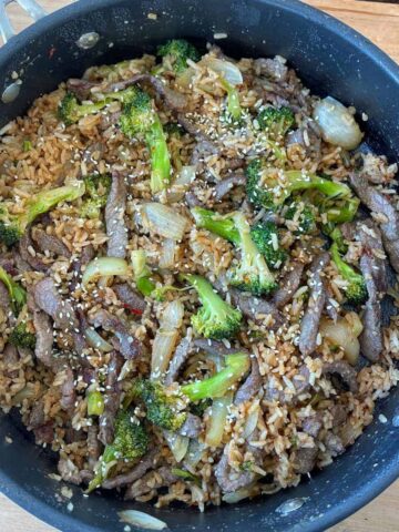 A skillet filled with beef stir fry, broccoli, onions, and rice, topped with sesame seeds.