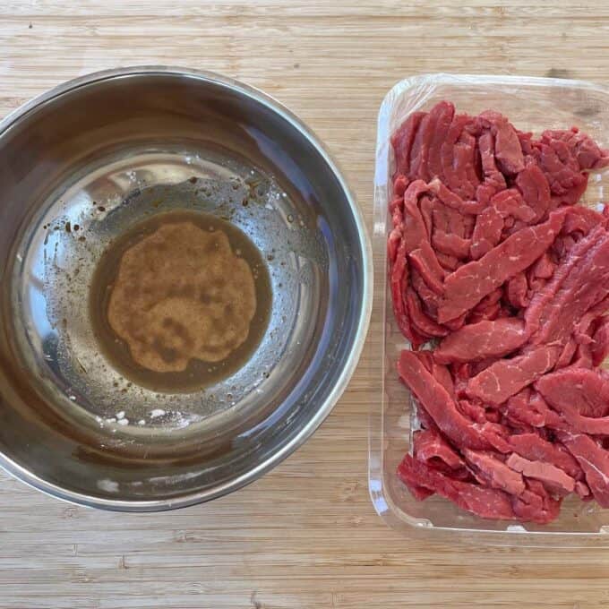 Beef and Broccoli Fried Rice A metal bowl with marinade next to a plastic container of raw beef strips on a wooden surface.