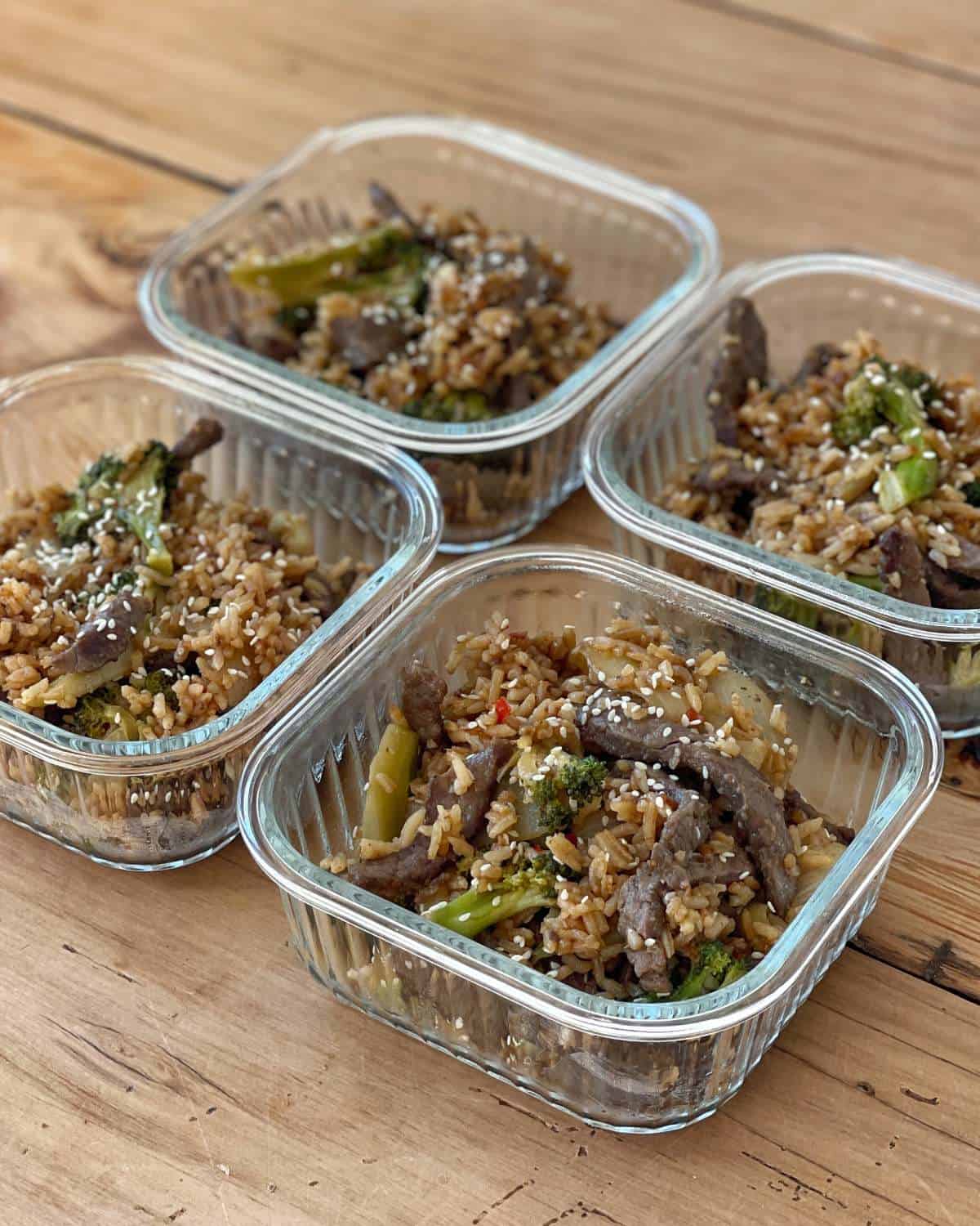 Beef and Broccoli Fried Rice Four glass containers filled with beef, broccoli, and rice stir fry are arranged on a wooden surface.