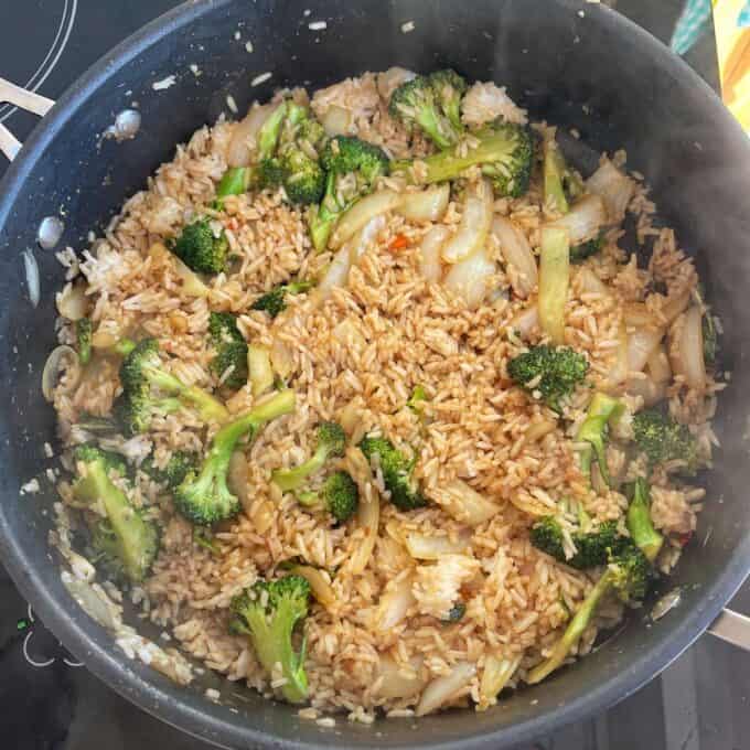 Beef and Broccoli Fried Rice A large pan filled with fried rice, broccoli florets, and onions, all mixed together and cooked on a stovetop.