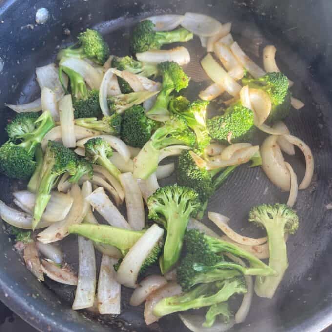 Beef and Broccoli Fried Rice Broccoli florets and sliced onions are being sautéed in a black frying pan, with light steam rising from the vegetables.