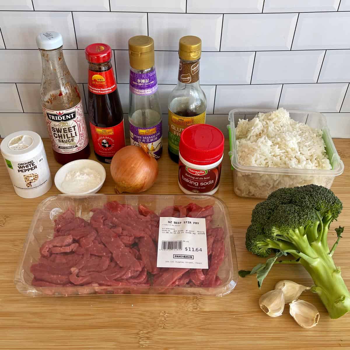 Beef and Broccoli Fried Rice Ingredients for a beef stir-fry arranged on a counter: sliced beef, broccoli, rice, sauces, onion, garlic, baking soda, pepper, and cornstarch in front of a white tile backsplash.