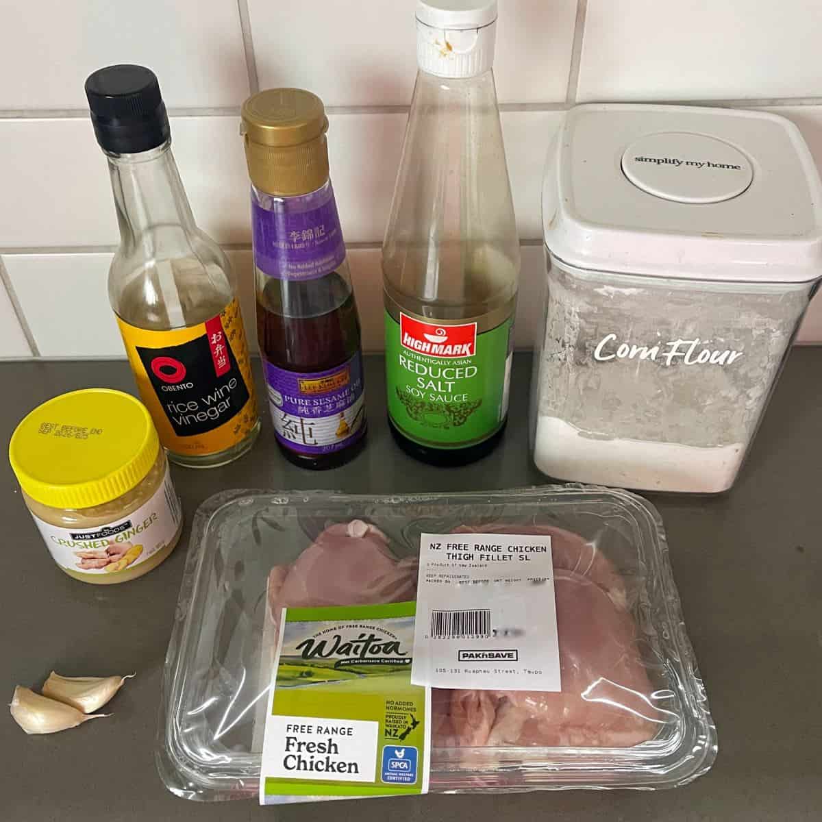 Air Fryer Karaage Chicken Raw chicken breast, garlic cloves, cornflour, rice wine vinegar, pure sesame oil, soy sauce, and a container of yellow paste are arranged on a kitchen counter.