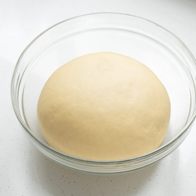 Biscoff Cinnamon Scrolls A ball of dough resting in a clear glass bowl on a white countertop.