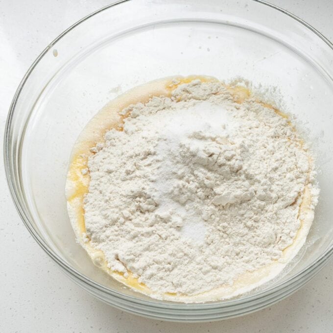 Biscoff Cinnamon Scrolls A glass bowl containing flour, sugar, and wet ingredients ready to be mixed on a white countertop.