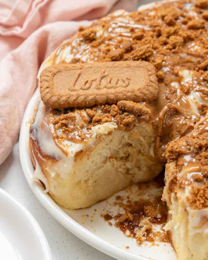 Biscoff Cinnamon Scrolls A close-up of a frosted cinnamon roll topped with Biscoff cookie spread, cookie crumbs, and a whole Lotus biscuit, with a slice missing from the roll.
