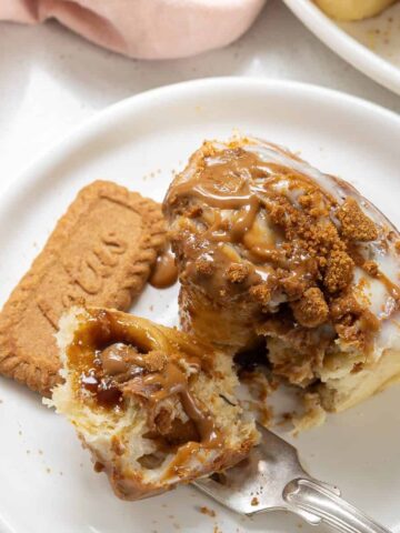 A cinnamon roll topped with icing and crumbled cookie sits on a white plate next to a whole cookie, with a fork cutting into the roll.