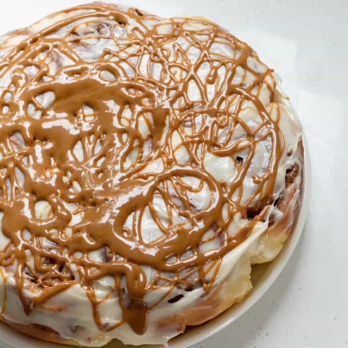 Biscoff Cinnamon Scrolls A round cinnamon roll cake covered with white icing, drizzled generously with brown caramel or caramel-flavored icing, on a white plate.