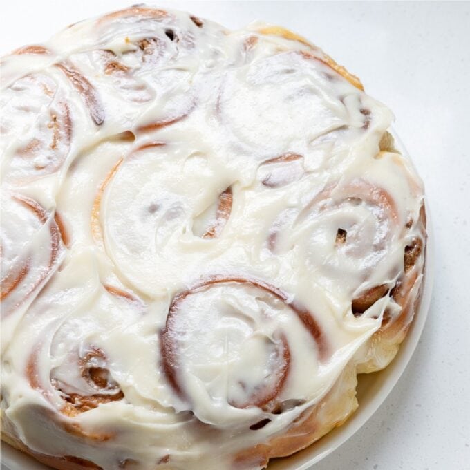 Biscoff Cinnamon Scrolls A plate of cinnamon rolls covered in white cream cheese icing, viewed from above on a white surface.