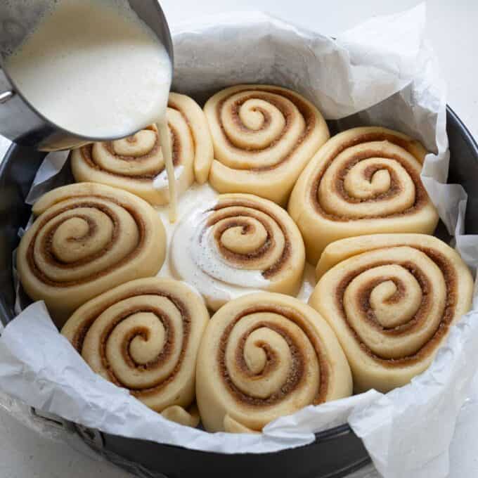 Biscoff Cinnamon Scrolls A person pours cream over unbaked cinnamon rolls arranged in a round, parchment-lined baking pan.