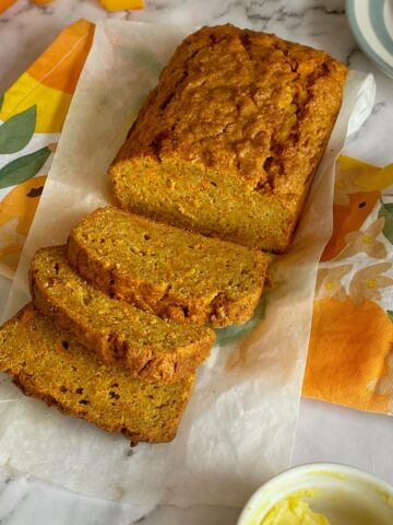 A loaf of sliced carrot bread on parchment paper, with a colorful floral napkin and a small dish of butter nearby.