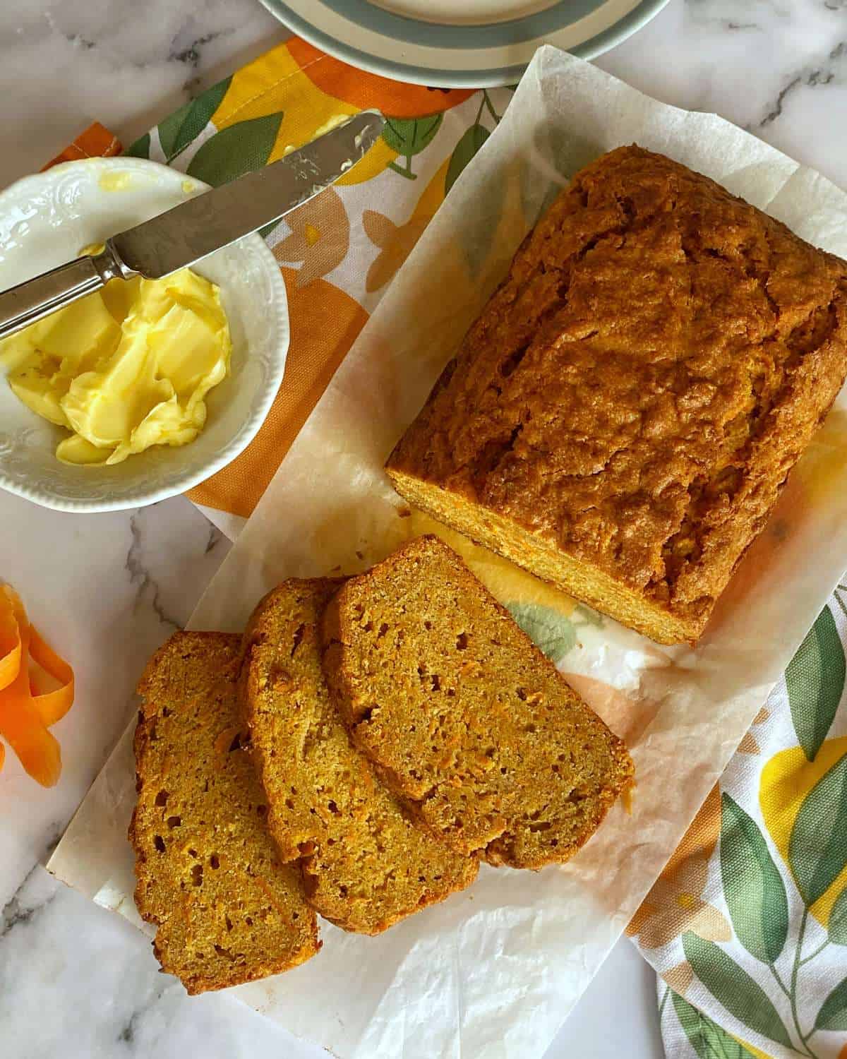A loaf of carrot bread sits on parchment paper, with three slices cut. A bowl of butter with a knife and a colorful napkin are beside it on a marble surface.