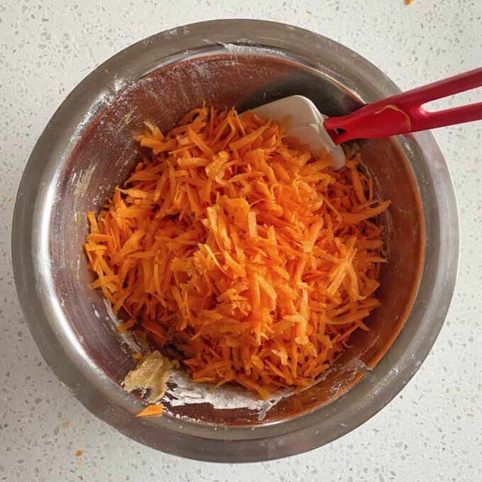 Carrot Cake Loaf A metal bowl with grated carrots, flour, and a red-handled spatula on a speckled countertop.