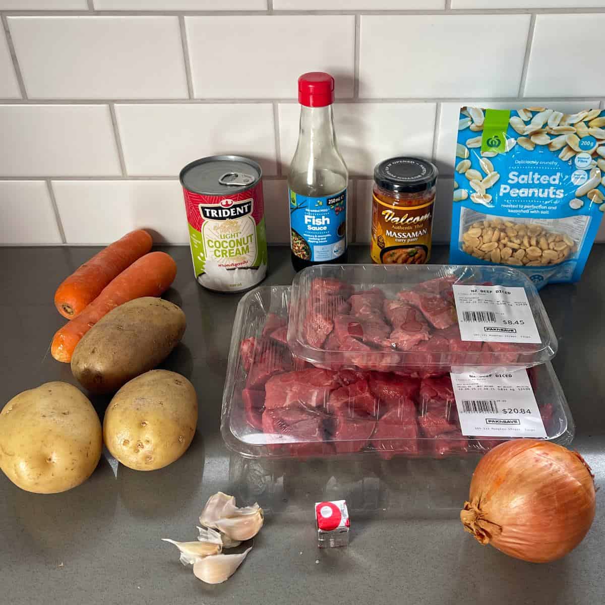 Massaman Beef Curry Ingredients for a meal on a countertop: carrots, potatoes, garlic, onion, beef cubes, coconut cream, fish sauce, massaman curry paste, salted peanuts, and a stock cube.