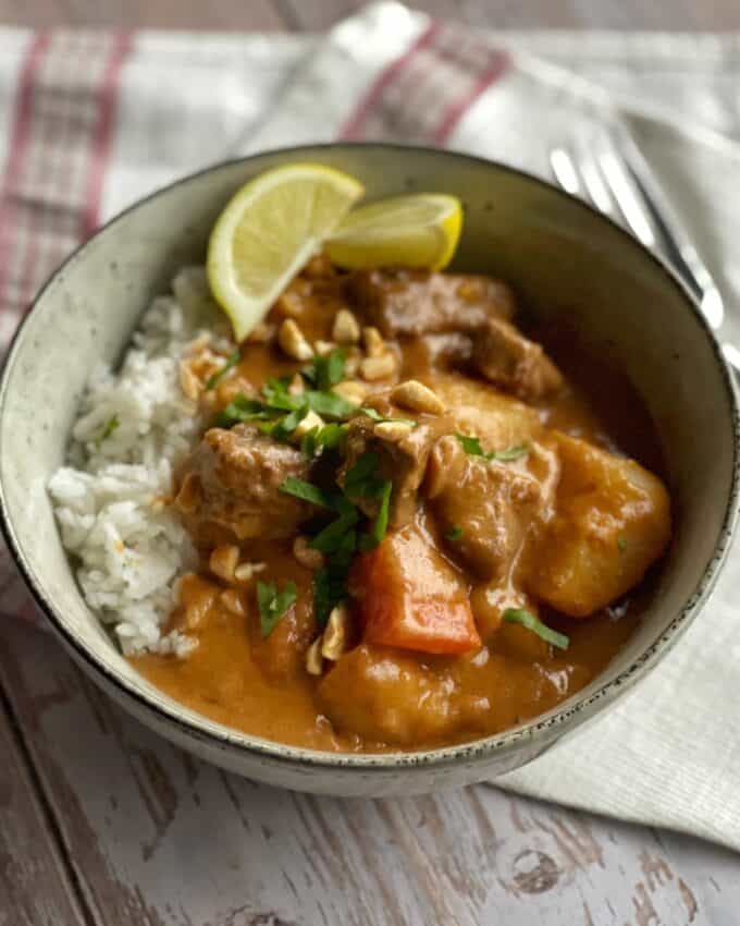 Massaman Beef Curry A bowl of white rice topped with beef stew in a reddish sauce, garnished with chopped herbs, peanuts, and lemon wedges, placed on a wooden table beside a fork and napkin.