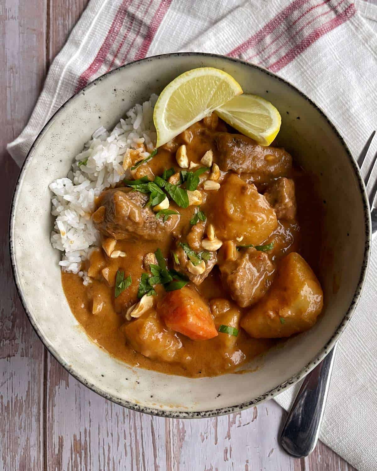 Massaman Beef Curry A bowl of rice topped with curry containing beef, potatoes, carrots, chopped herbs, and nuts, garnished with lemon wedges.