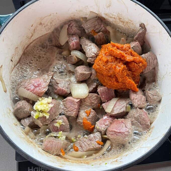 Massaman Beef Curry Chunks of beef and onions cooking in a pot with a dollop of red paste and minced garlic on top.