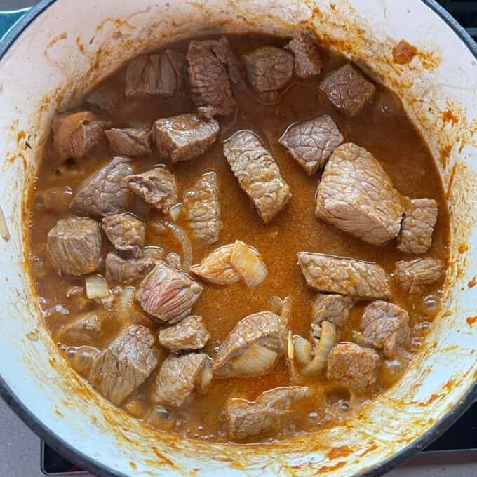Massaman Beef Curry Chunks of beef simmering in a reddish-brown sauce with pieces of onion in a large pot on a stovetop.