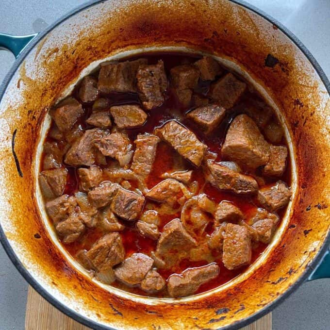 Massaman Beef Curry Chunks of beef simmering in a red sauce inside a stained, enameled cast iron pot on a countertop.