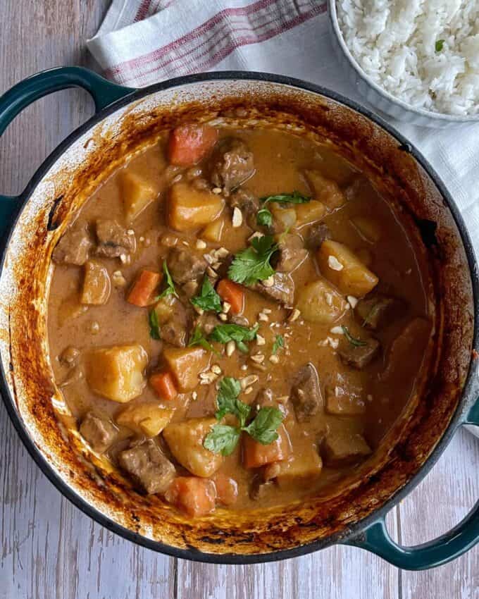 Massaman Beef Curry A pot of beef stew with potatoes, carrots, and fresh cilantro garnish, next to a bowl of white rice on a wooden table.