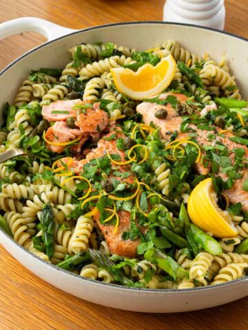 A white pan filled with rotini pasta, salmon fillets, lemon wedges, capers, and chopped herbs, garnished with lemon zest.