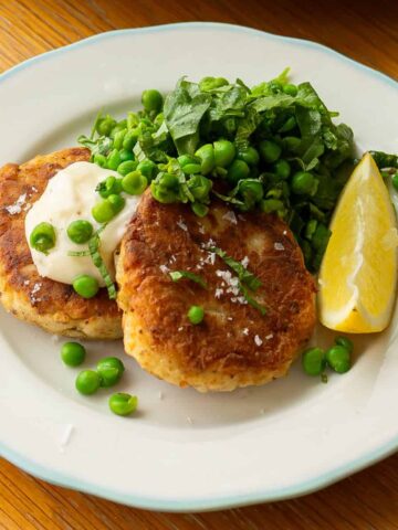 Two fish cakes served with a dollop of mayonnaise, green peas, chopped greens, and a lemon wedge on a white plate.