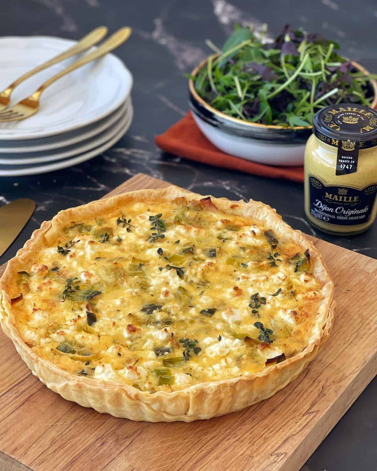 A baked vegetable quiche on a wooden board, with a bowl of mixed greens, a jar of Dijon mustard, and stacked plates with cutlery in the background.