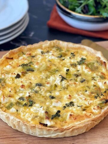 A baked quiche with a golden crust and visible herbs sits on a wooden board, with plates and a bowl of salad in the background.